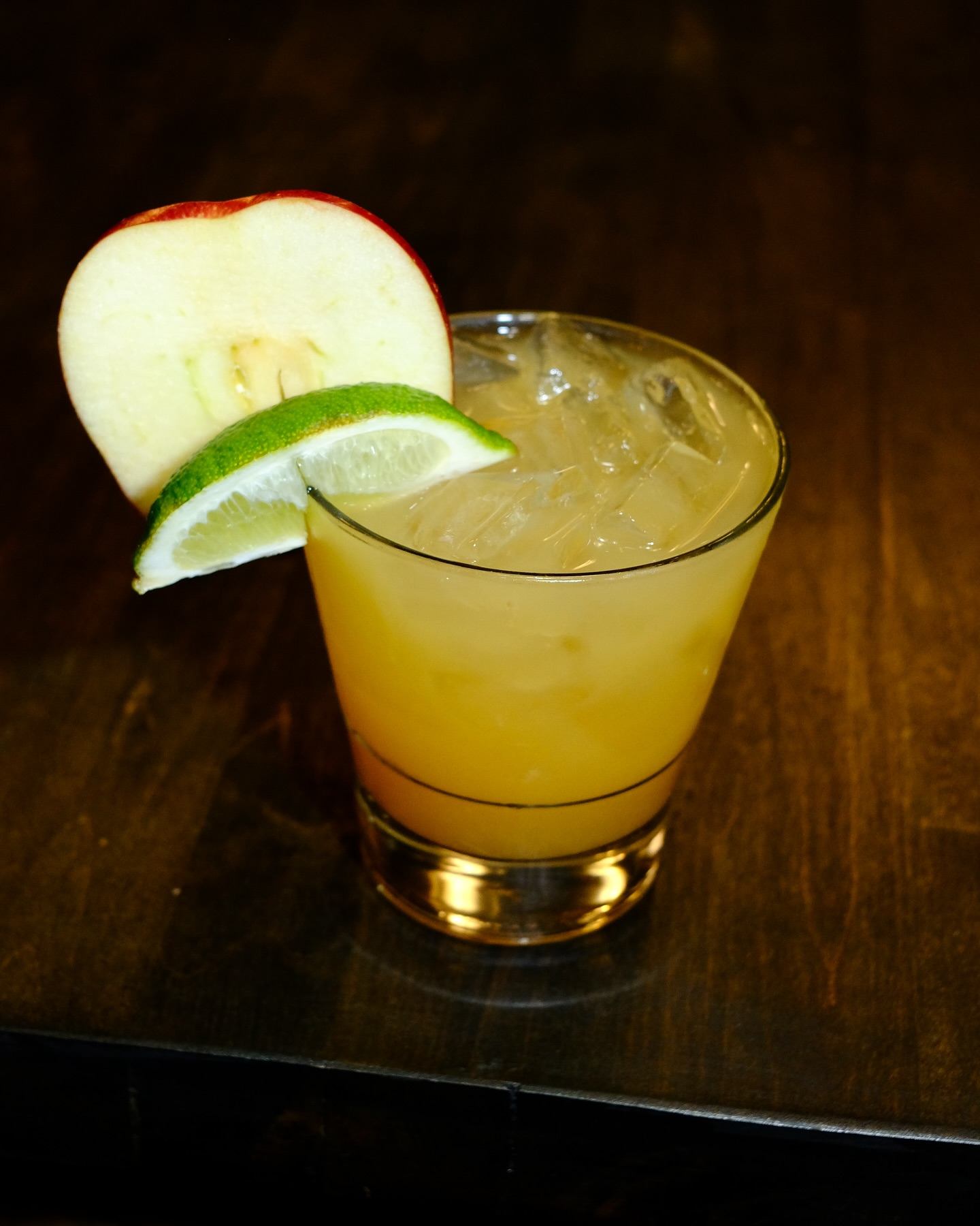 Glass of yellow iced drink with lime and apple garnishes.