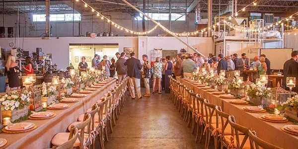 People socialize between long decorated tables in an industrial-style event space.