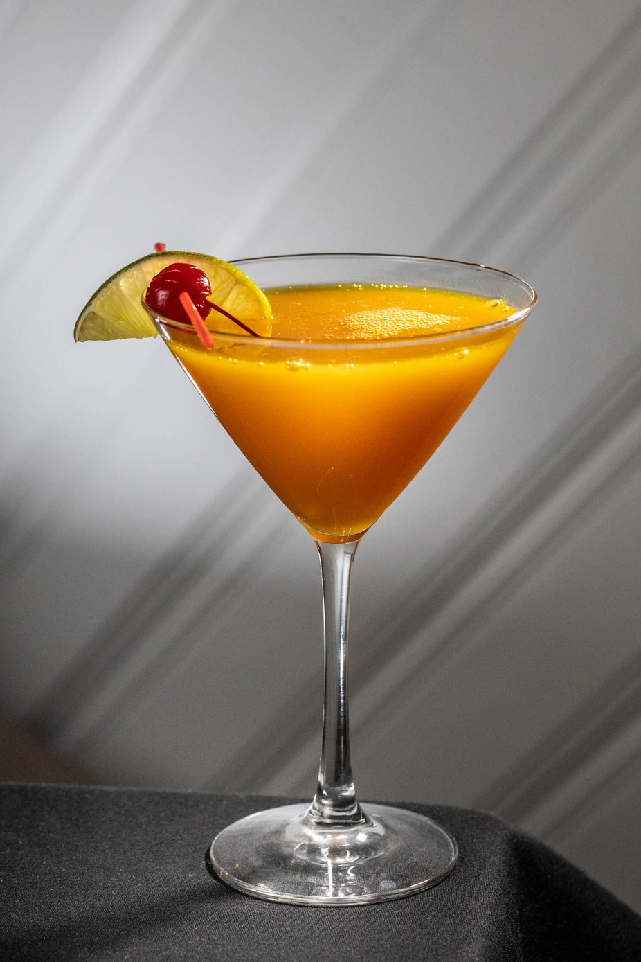Orange cocktail in martini glass, garnished with lime and cherry.