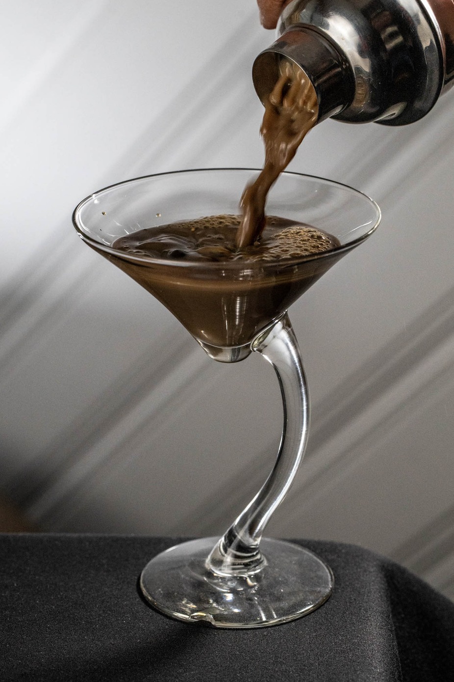 Brown liquid poured from shaker into martini glass with curved stem.