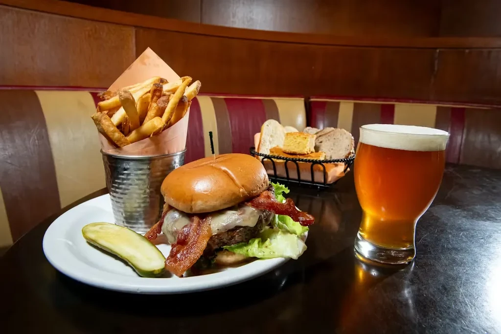 Bacon cheeseburger, fries, pickle, and beer on a table.