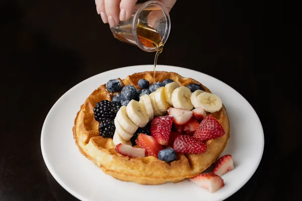 Maple syrup pouring over waffle with blueberries, blackberries, bananas, and strawberries.