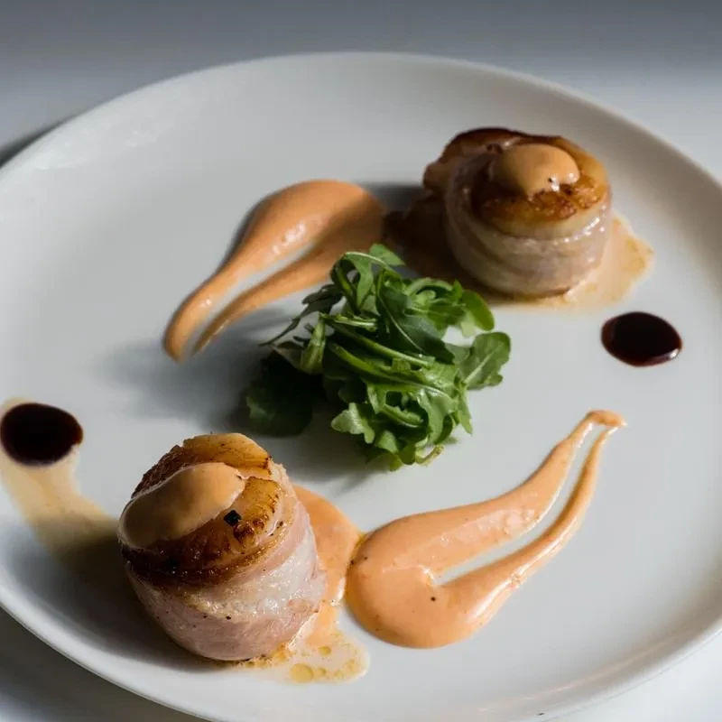 Two bacon-wrapped scallops with sauce and greens on a white plate.