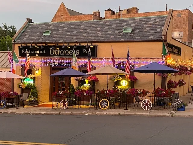 Dunne's Pub exterior with outdoor seating, flags, and string lights at dusk.