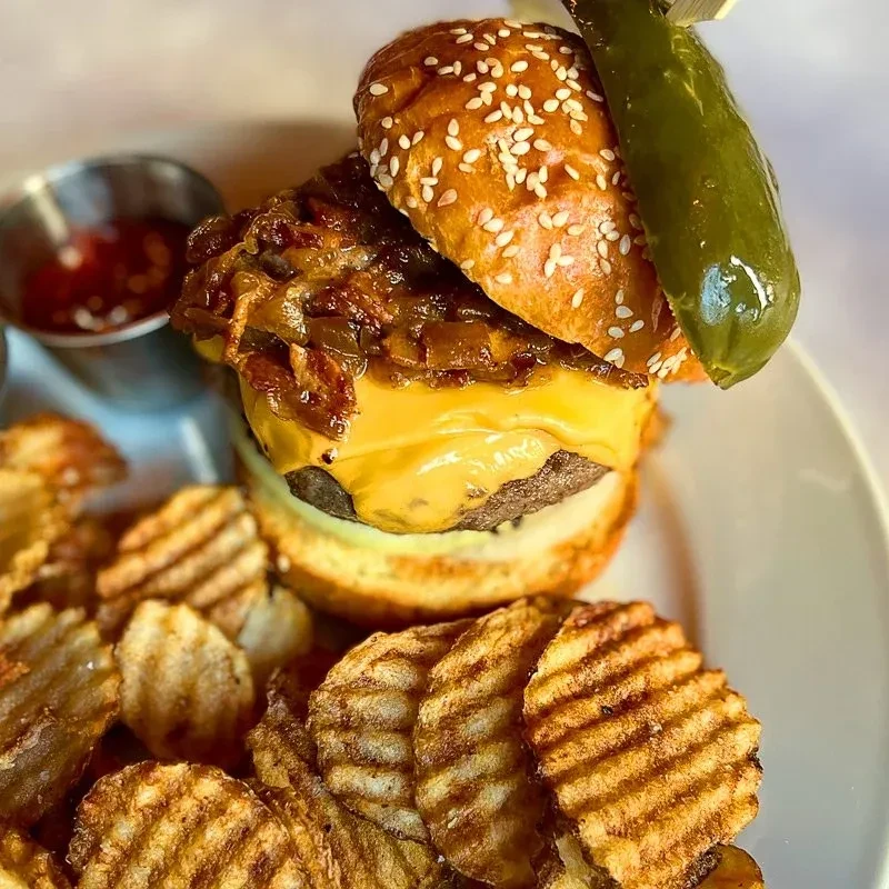 A hamburger with caramelized onions, melted cheese, waffle chips, and a pickle spear.