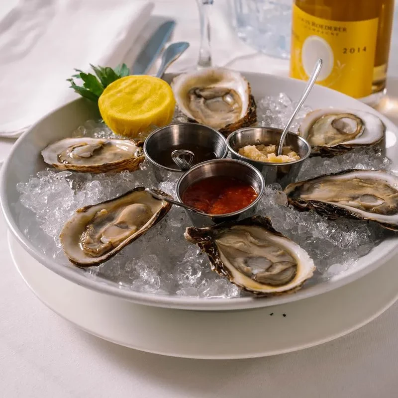 A white bowl of oysters on ice with sauces and lemon.