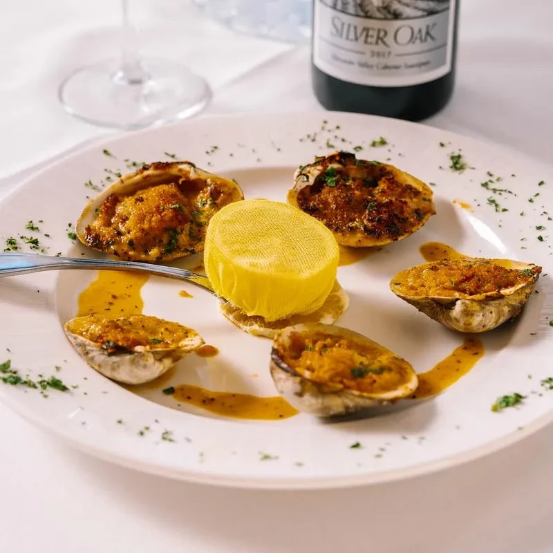 A white plate with six baked clams, lemon, and a wine bottle.