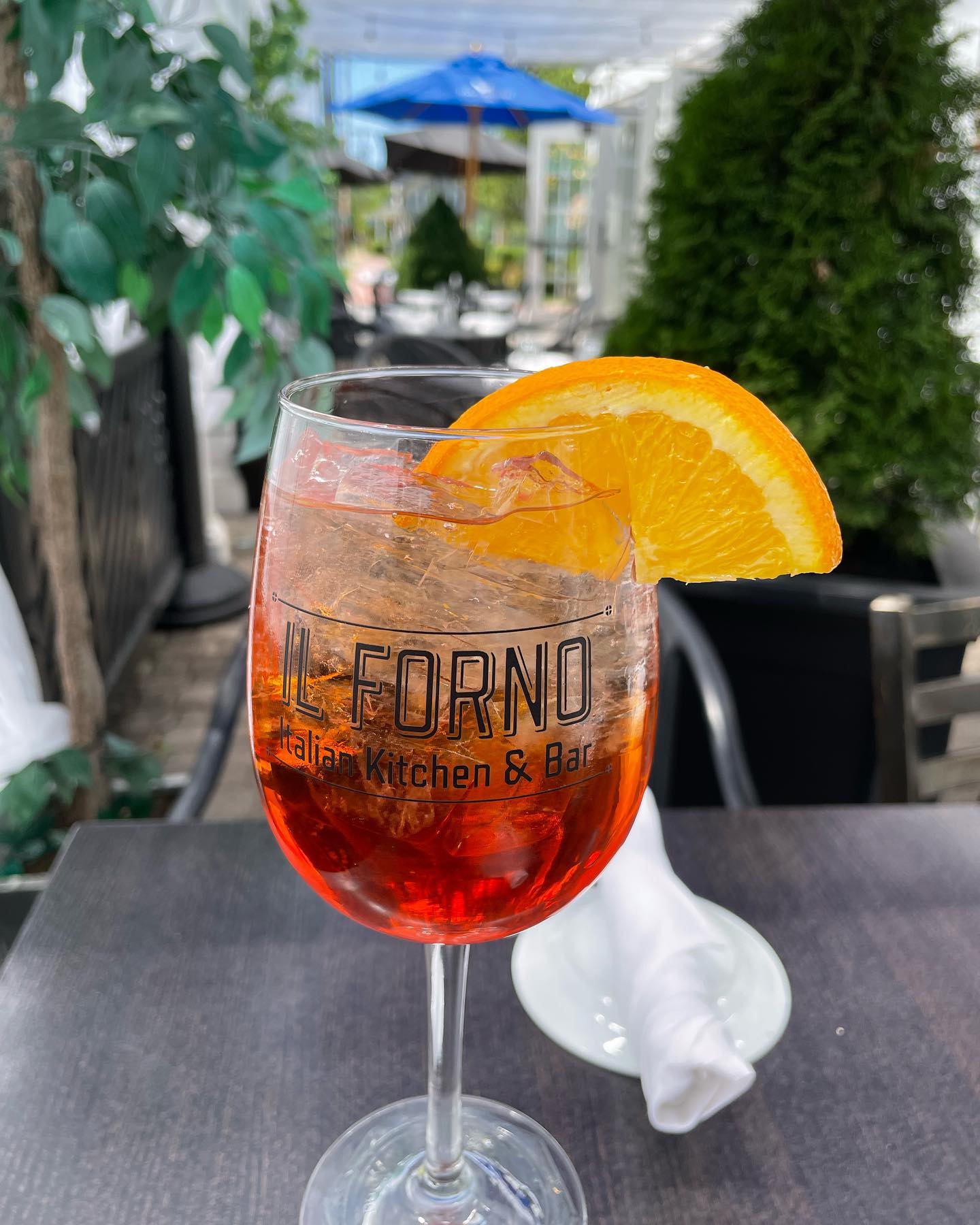 Reddish-orange drink with ice, orange slice, and 'IL FORNO' glass.