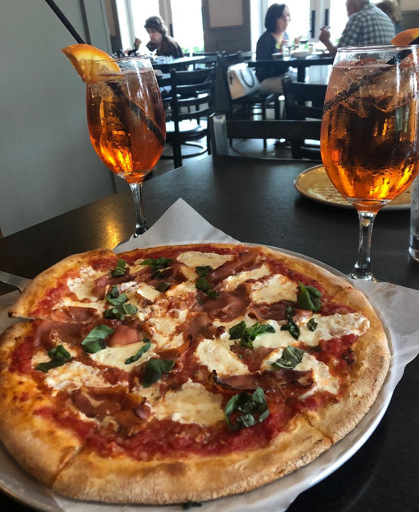 Pizza with prosciutto, basil, and two orange cocktails on a restaurant table.