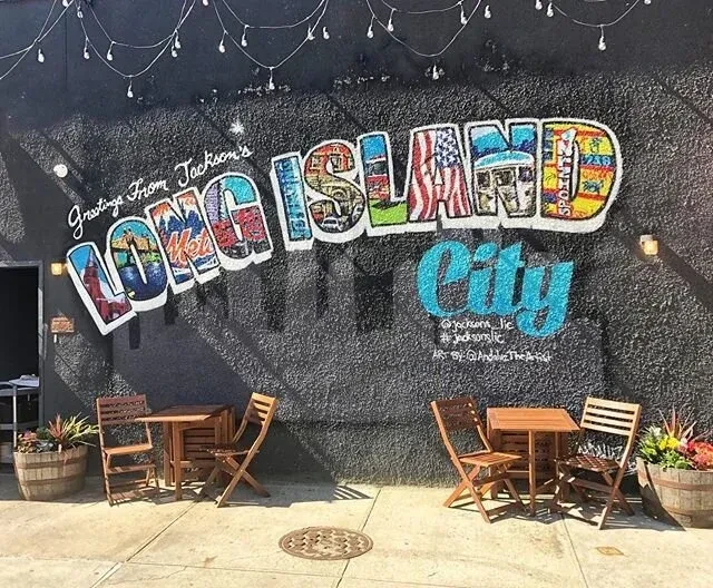 Mural with "Greetings from Long Island City" on a black wall with outdoor tables.