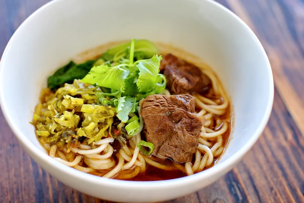 White bowl of beef noodle soup with greens and pickled vegetables.