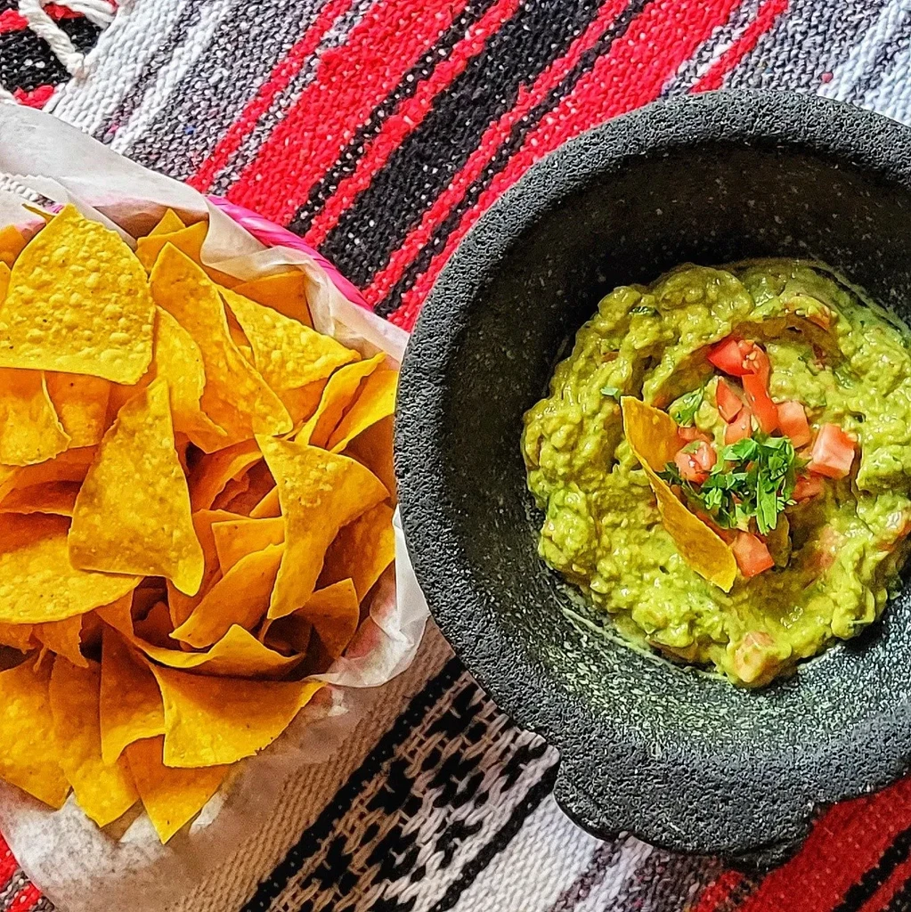 A bowl of guacamole next to a basket of tortilla chips.