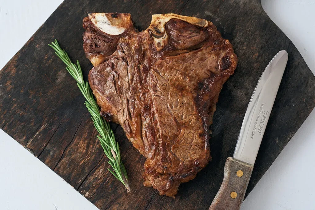 Cooked T-bone steak, rosemary, and knife on a wooden board.