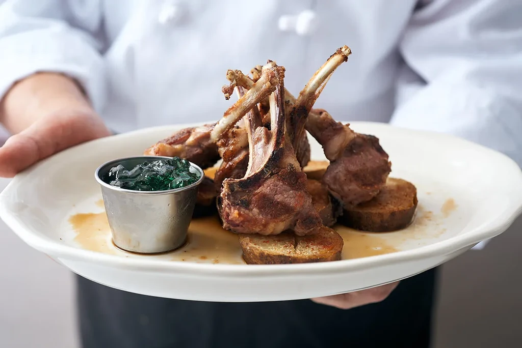 A chef holding a plate of lamb chops, potatoes, and mint jelly.