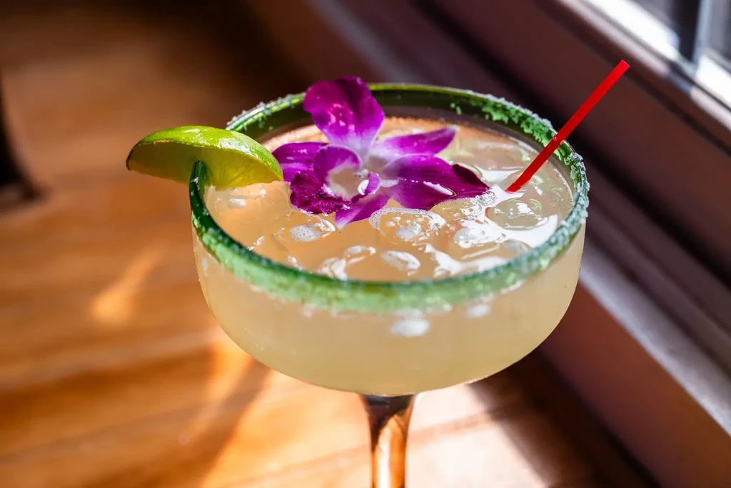 Margarita with lime, purple orchid, and red straw on a table.