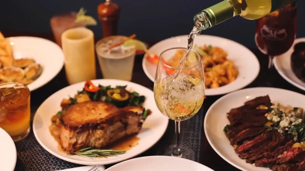 Pouring white wine into a glass surrounded by plates of food.