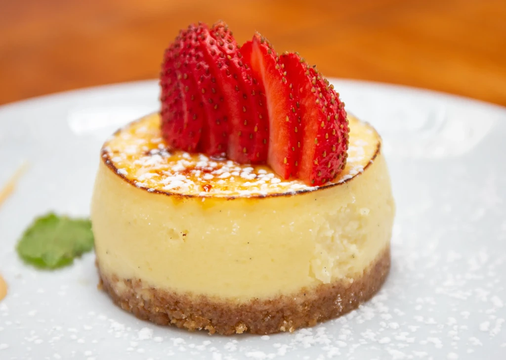 A mini cheesecake with sliced strawberries, caramelized sugar, and powdered sugar.