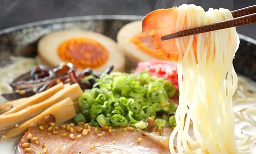 Chopsticks lifting steaming ramen noodles with pork, eggs, and green onions.