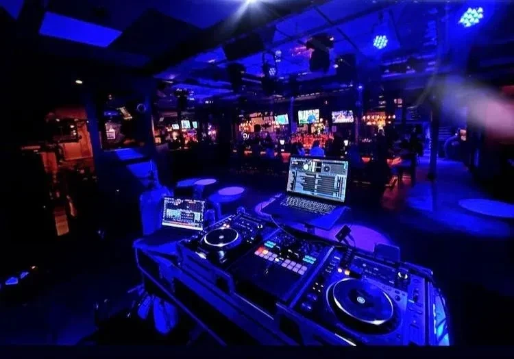From behind a DJ setup, a blue-lit nightclub with a bar.
