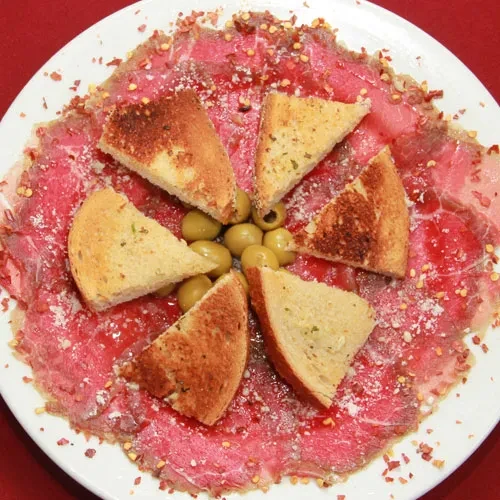 Overhead carpaccio dish with olives, cheese, chili flakes, and toasted bread.