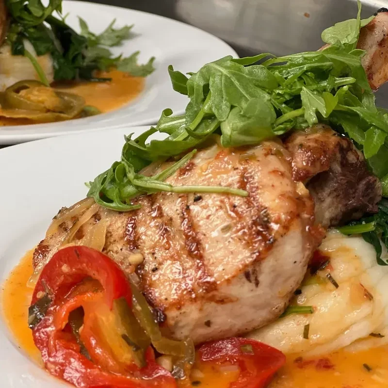 Grilled pork chop, mashed potatoes, roasted peppers, and arugula on a plate.