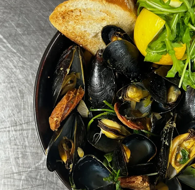 A black bowl of mussels, bread, and lemon on a metal surface.