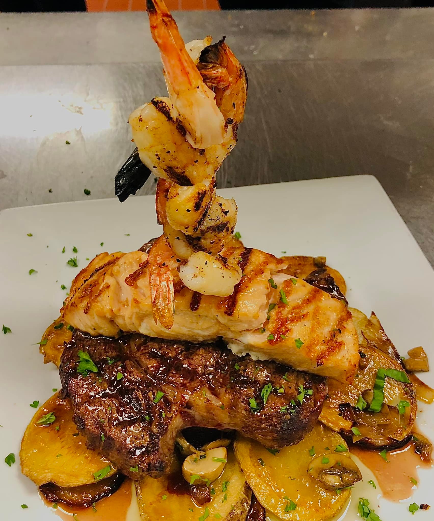 Stacked grilled shrimp, salmon, steak, potatoes, mushrooms, garnished with parsley.