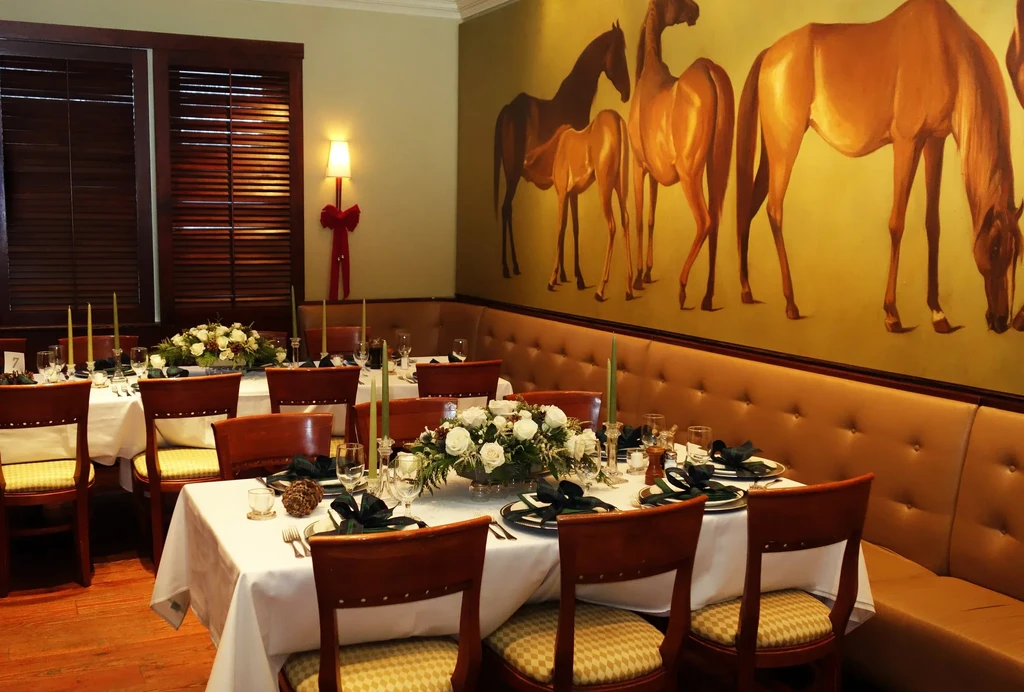 Formal dining room with white-clothed tables, horse mural, and festive sconce.