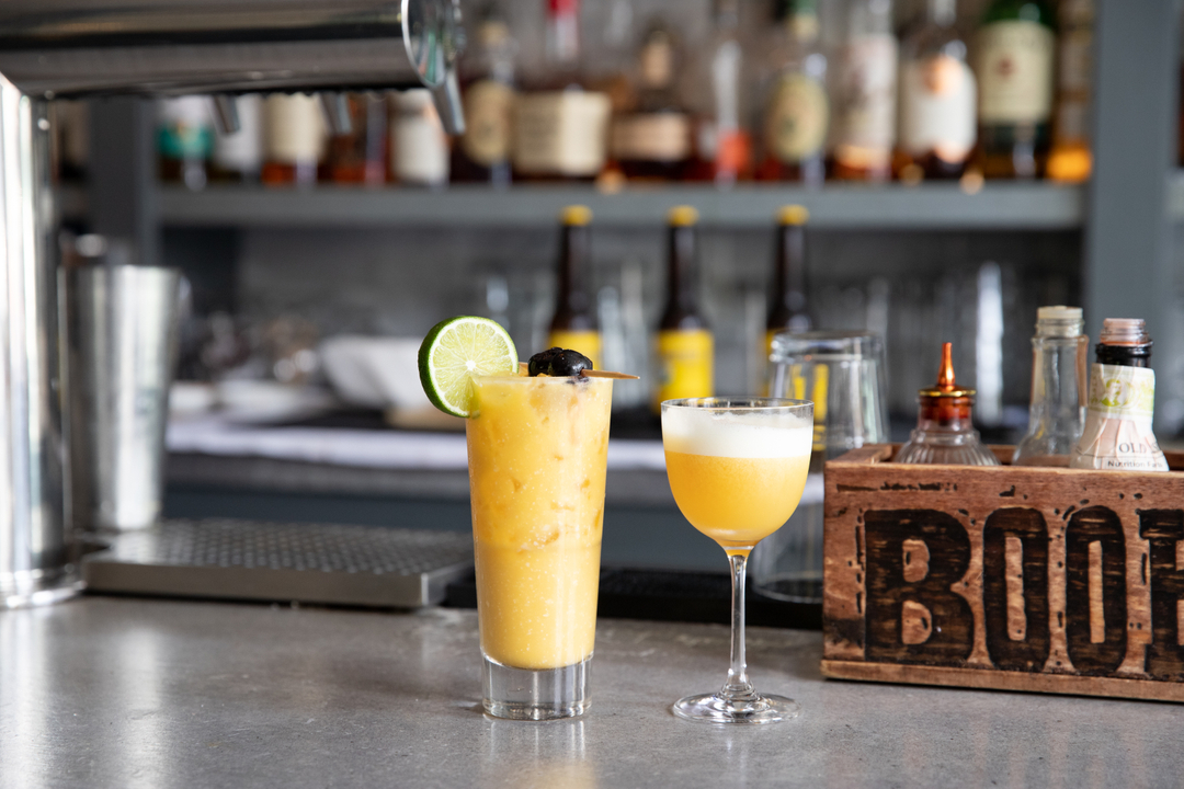 Two cocktails on a bar counter with bottles in the background.