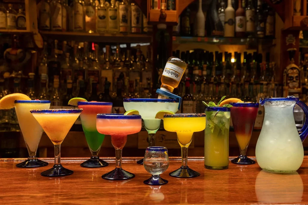 Nine colorful margaritas and cocktails, one with beer, on a wooden bar.