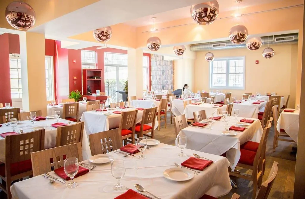 A brightly lit restaurant dining room with many set tables and red chairs.