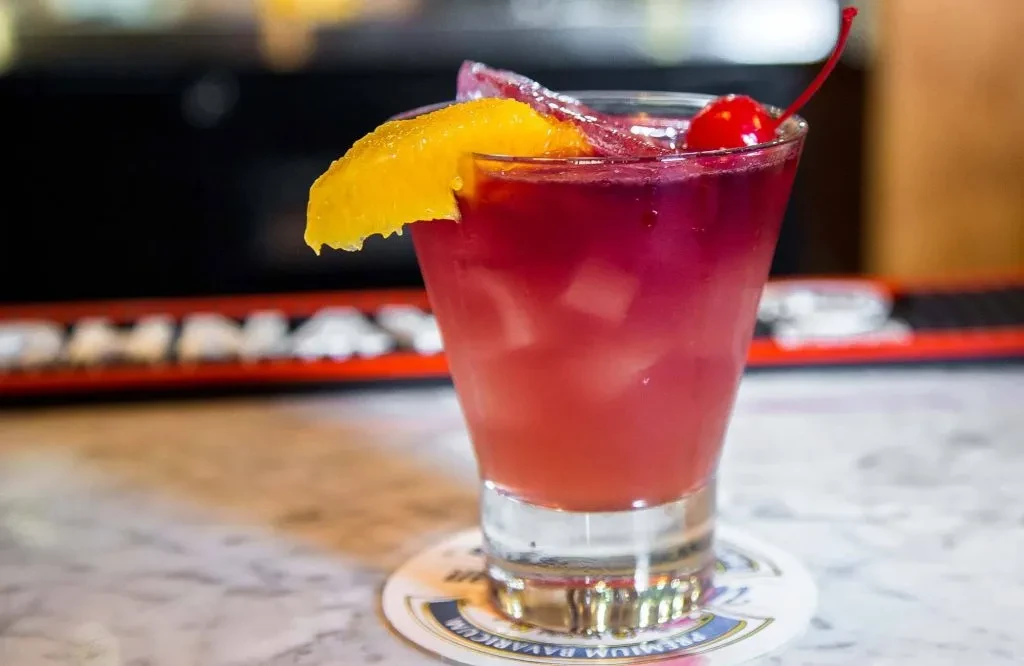 A red and purple cocktail with a maraschino cherry and orange peel.