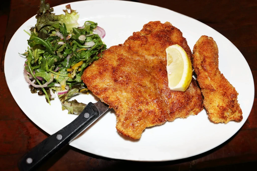 Crispy fried pork schnitzel, salad, and a lemon wedge on a white plate.