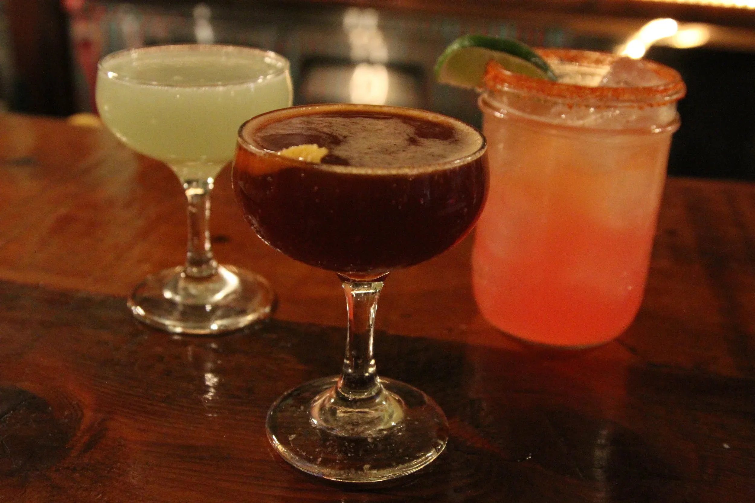 Three cocktails: pale green, dark brown, and coral.