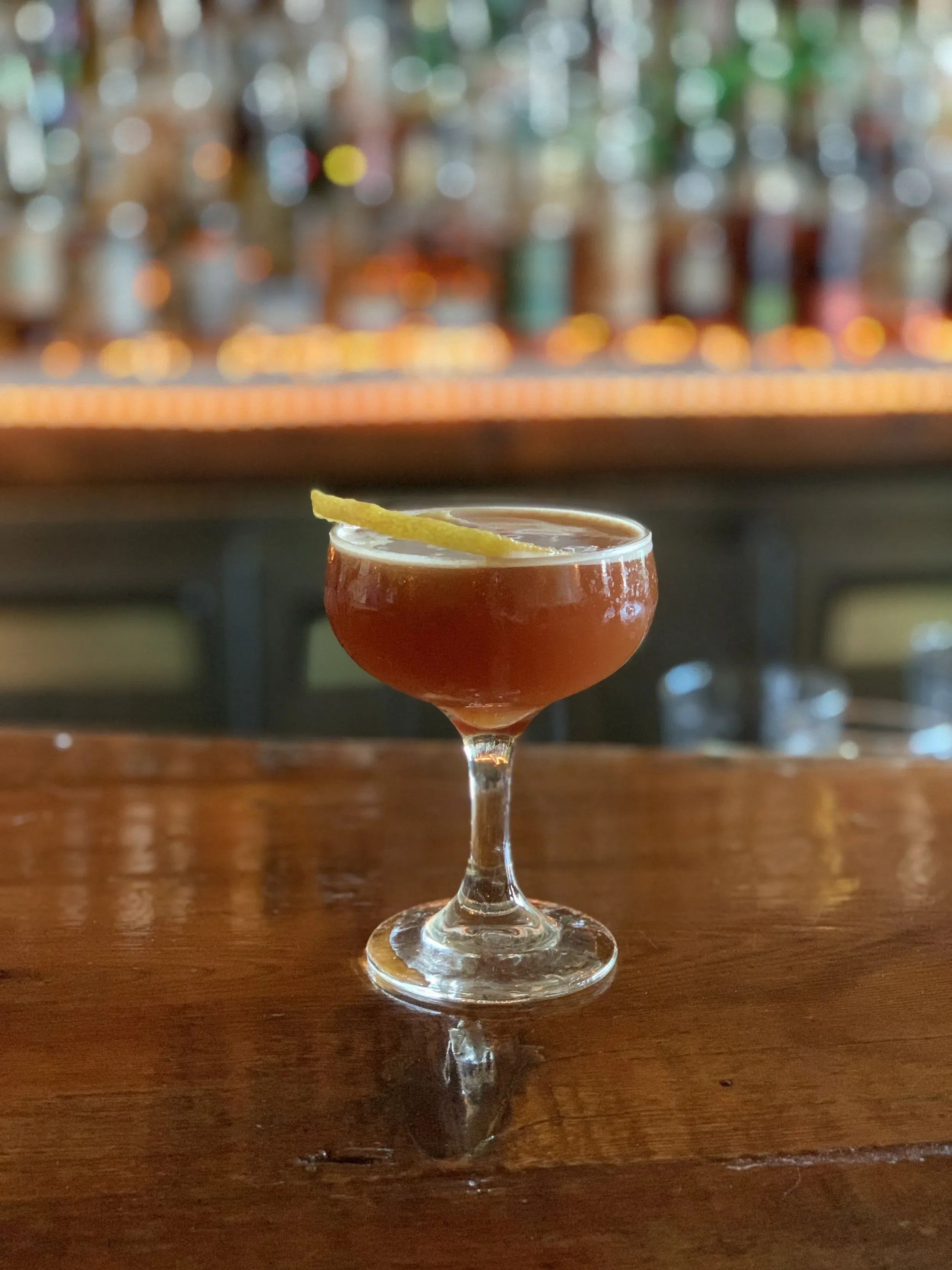 A coupe glass with an amber cocktail and a lemon peel on a wooden bar.