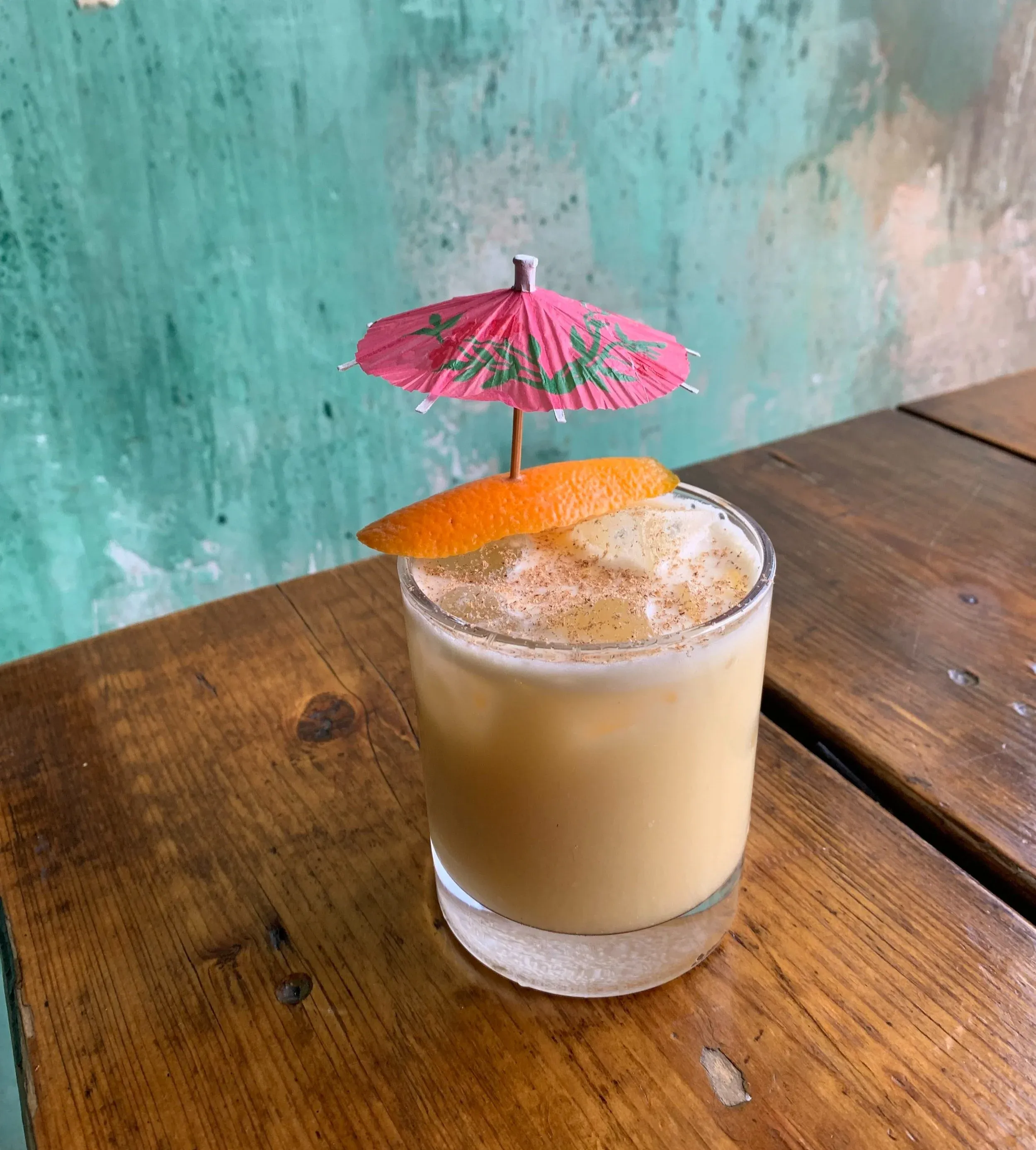 Creamy beverage with orange peel and paper umbrella on a wooden table.