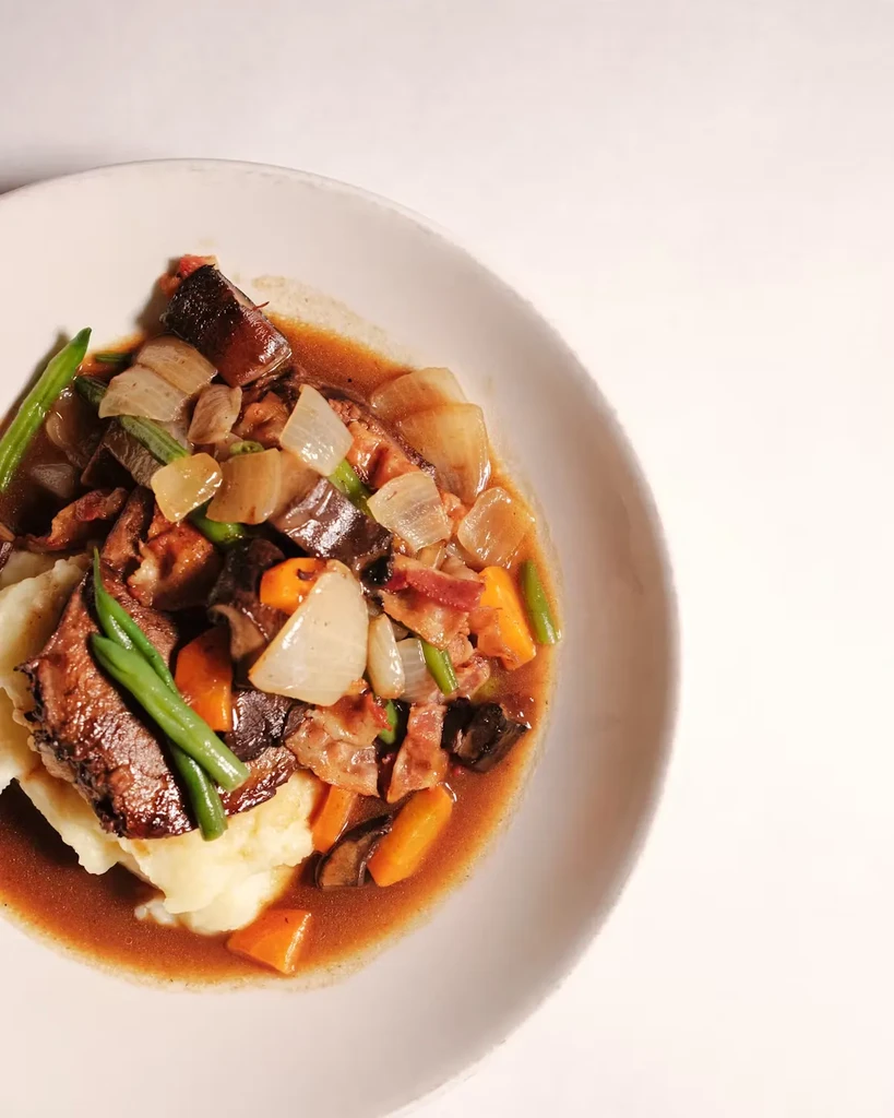 Braised short ribs with mashed potatoes, vegetables, and sauce in a bowl.