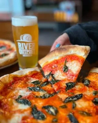 A hand lifts a slice of pizza with a beer and another pizza.