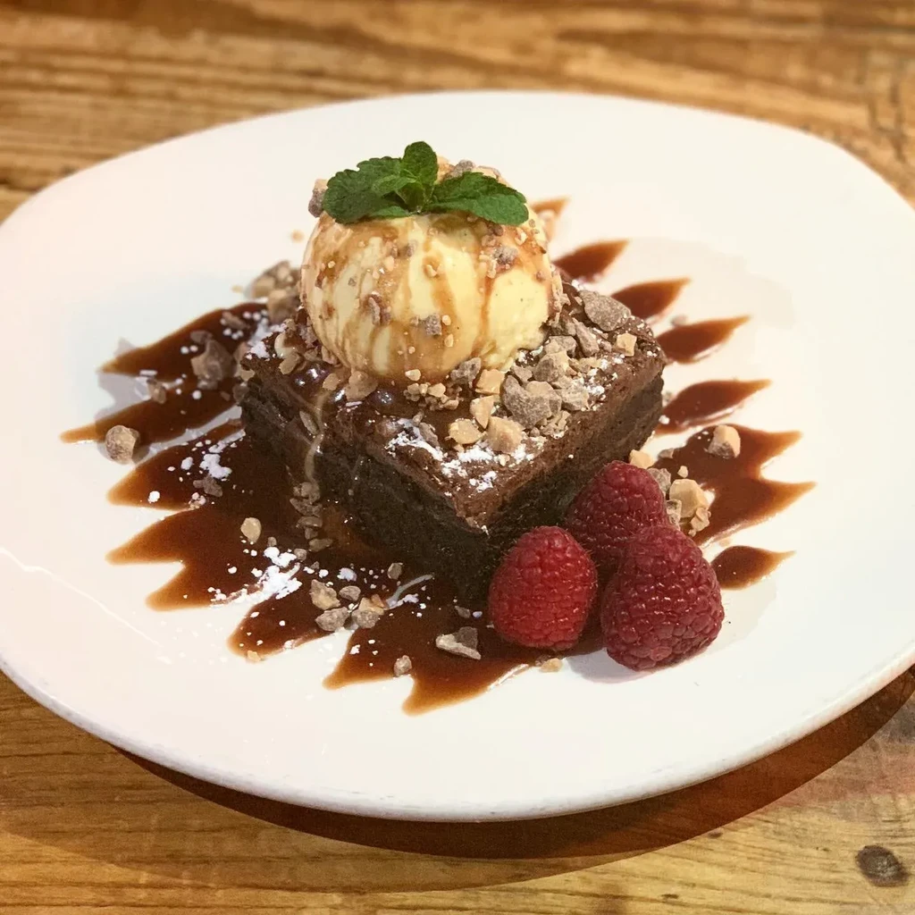 Brownie with ice cream, raspberries, caramel, and chopped nuts on a white plate.
