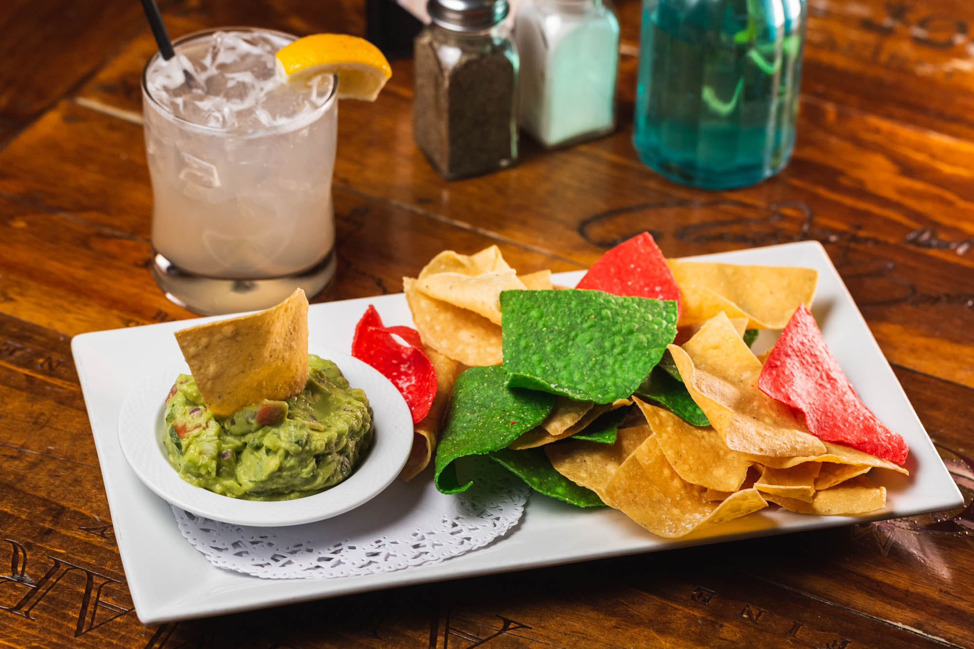 Guacamole, colorful tortilla chips, and an iced drink on a wooden table.