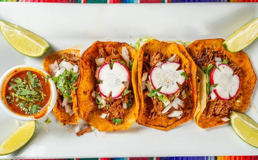 Four tacos with meat, radish, onion, cilantro, lime wedges, and dipping sauce.