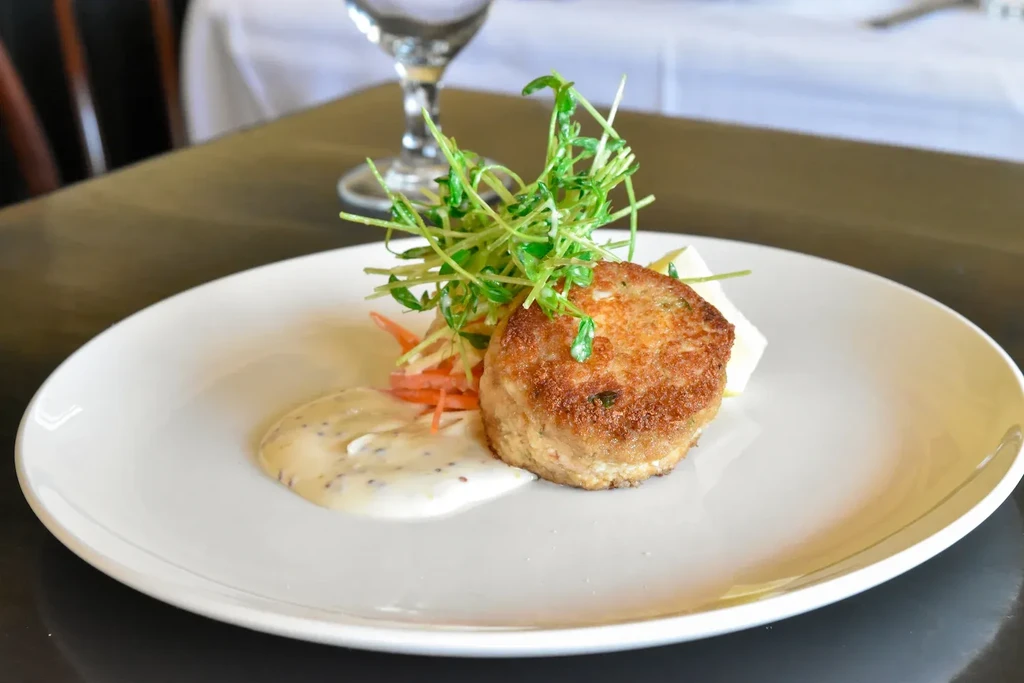A pan-fried crab cake with greens, sauce, and coleslaw on a white plate.