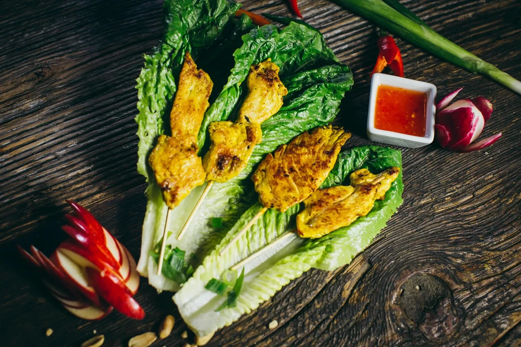Chicken satay skewers with lettuce, sauce, and radish on wooden table.
