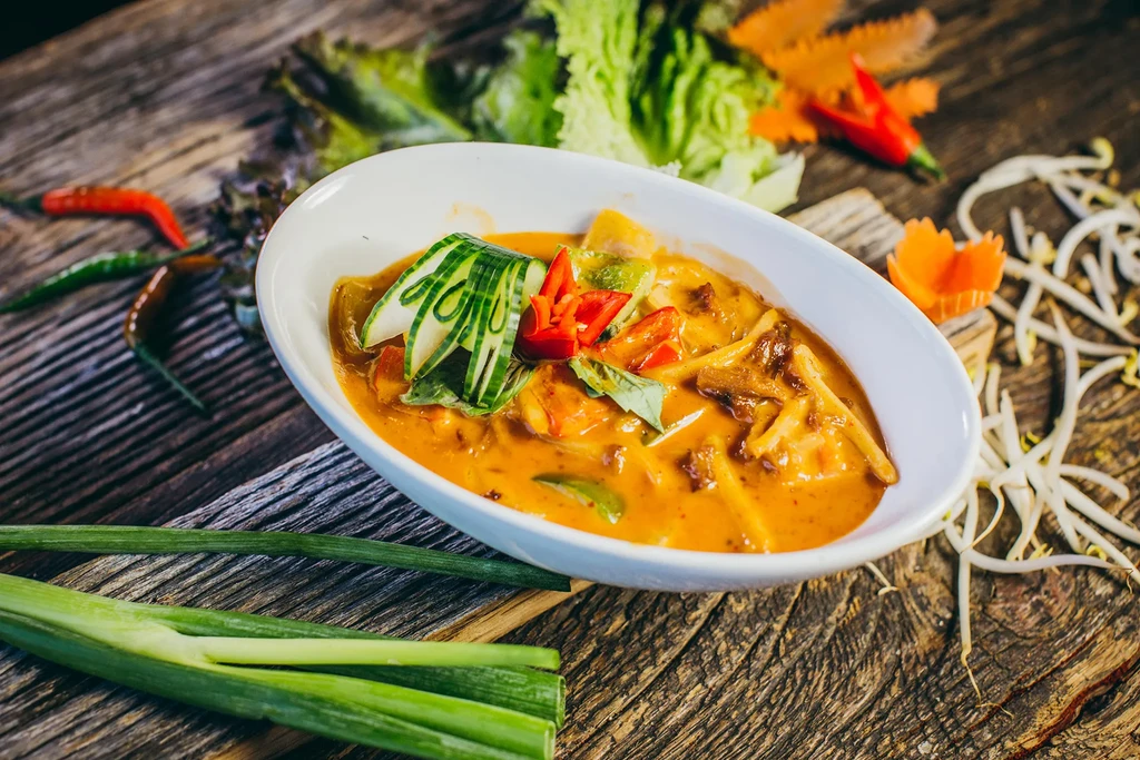 A white bowl of curry with vegetables and herbs on a wooden table.
