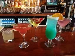 Five colorful cocktails on a wooden bar in front of liquor bottles.