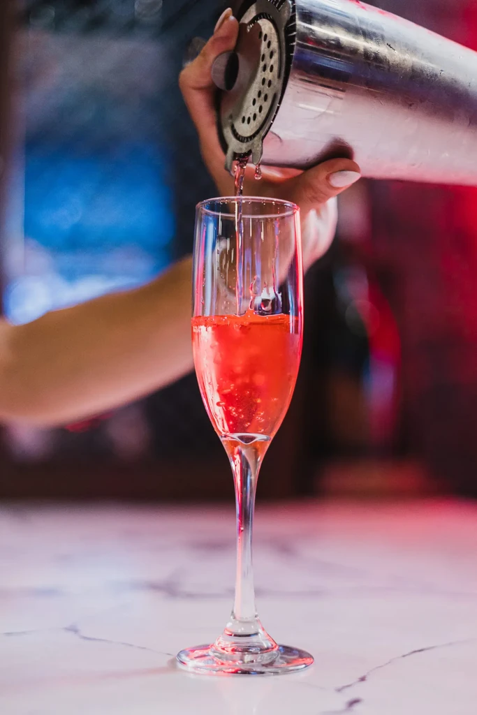 Someone pours a red cocktail from a shaker into a champagne flute.