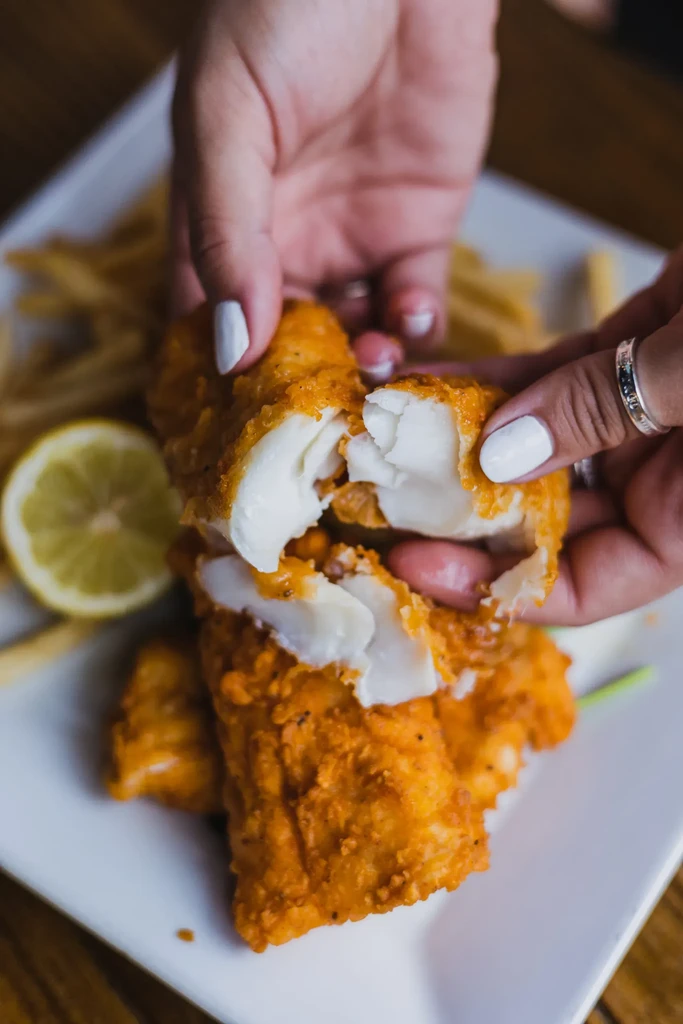 Hands with white nail polish pulling apart a piece of fried fish.