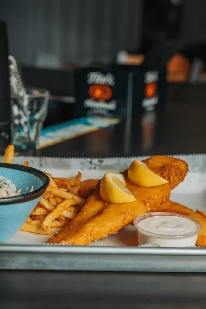 Fish and chips, coleslaw, and dipping sauce on a metal tray.
