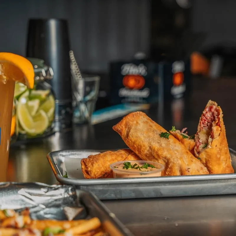 Golden-brown egg rolls with corned beef filling, sauce, and an amber drink.