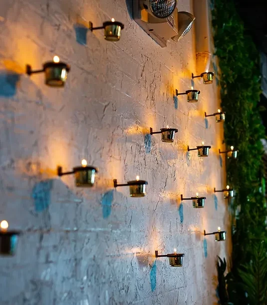 Textured wall displays staggered lit candles, dark holders, and green foliage.
